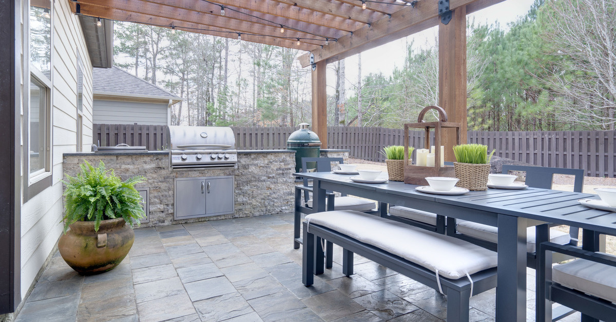 A spacious stone deck in a residential backyard. The shining sun peaks through a wooden pergola covering.